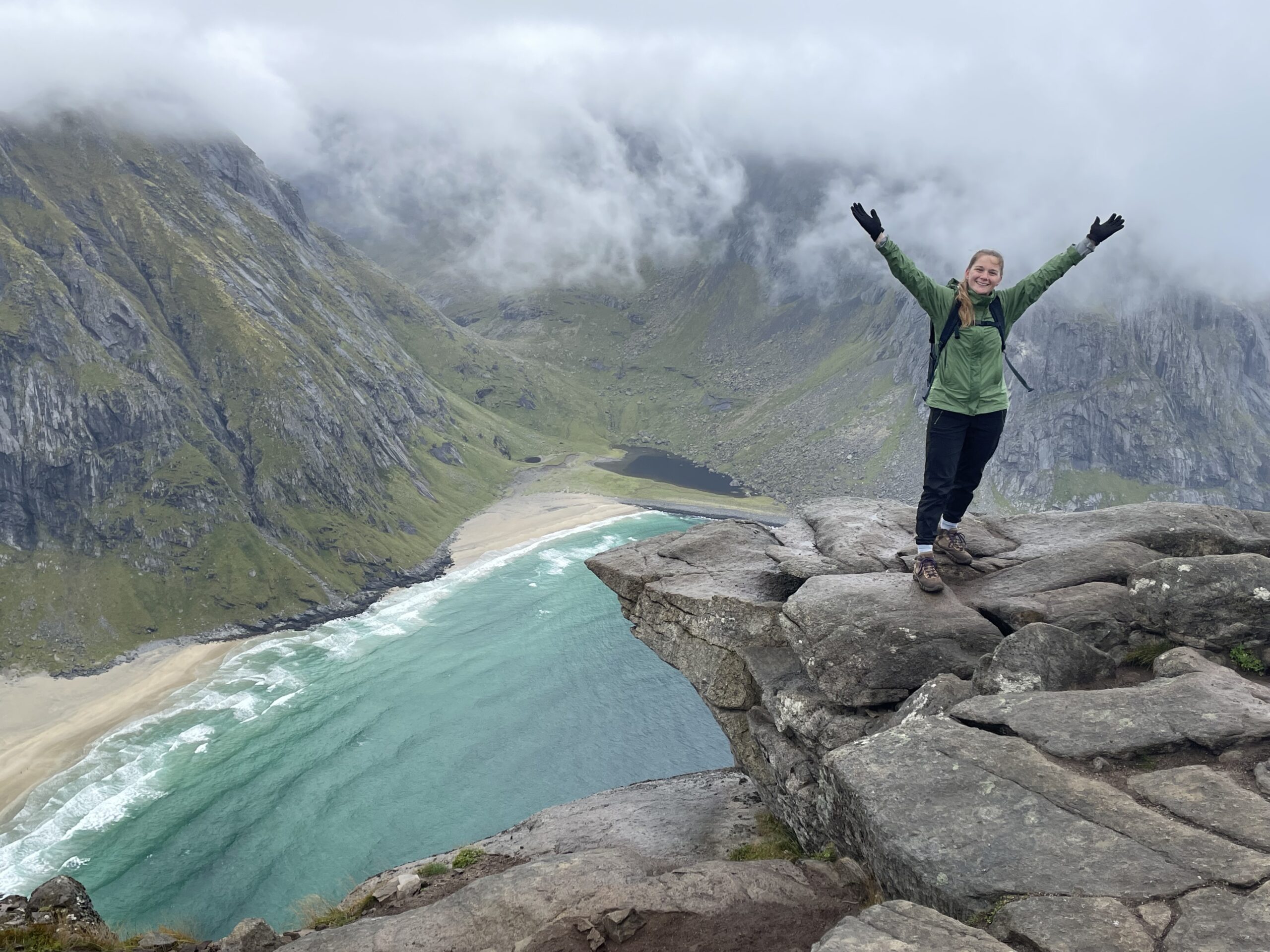 Janel standing on a cliff overlooking mountain with arms up